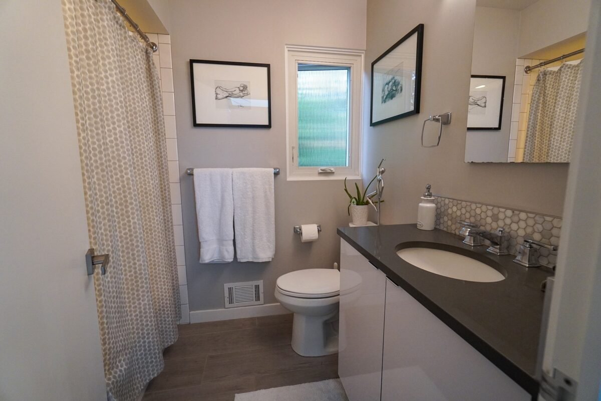 Bright, modern bathroom at Naakef Travel Nurse Stay – Lake Forest Park featuring a tiled shower-tub with patterned curtain, sleek dark countertop with under-mount sink, white vanity, artwork, and a frosted window for natural light and privacy.