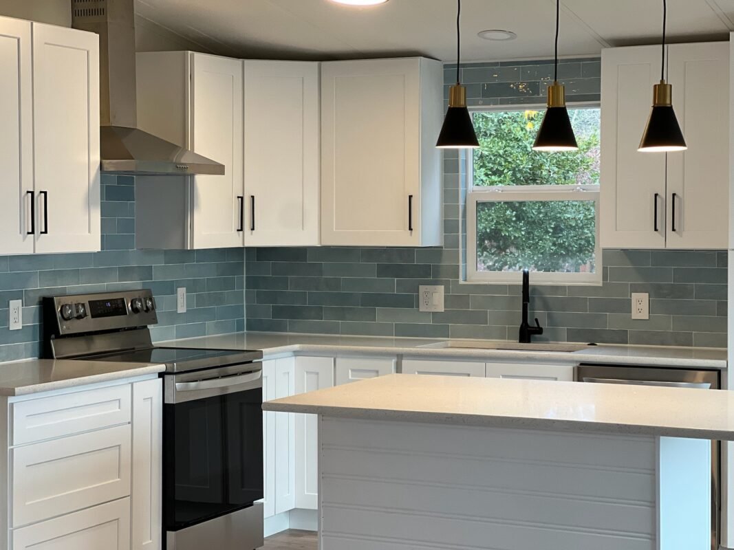 Bright open-concept kitchen at Naakef Private Home Rentals and RV Rentals – Seatac, featuring white cabinetry, a light blue kitchen island, teal subway tile backsplash, stainless steel appliances, and stylish black pendant lights.