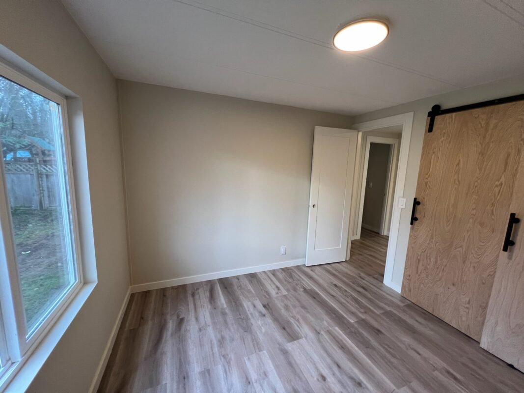 Spacious bedroom at Naakef Private Home Rentals and RV Rentals – Seatac with wood-look flooring, large window overlooking outdoor space, white doors, and a modern sliding barn-style closet door.
