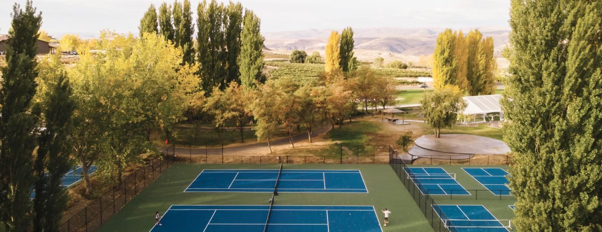 Aerial view of multiple blue and green pickleball and tennis courts at Sagecliffe Residences by Naakef surrounded by tall poplar trees and natural landscape.